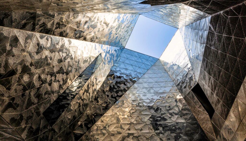A geometric architectural interior with reflective surfaces converging towards a bright blue sky opening above.