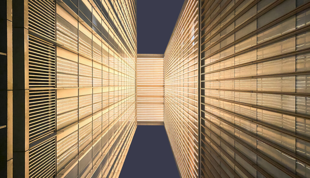 Looking up between two modern buildings, showcasing illuminated facades and sleek lines against a dark sky.