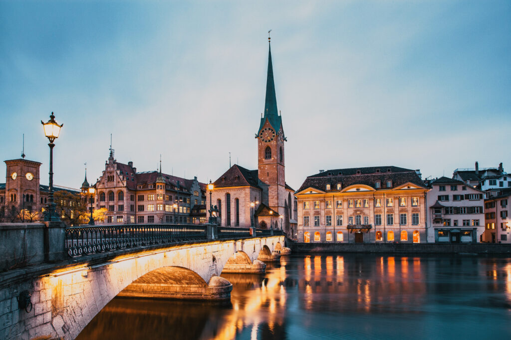 Twilight view of Zurich's skyline with a historic church, bridge, and lit buildings.