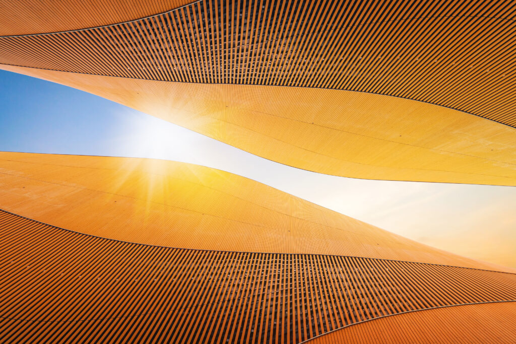 Abstract architectural view featuring curved, golden patterns against a bright blue sky with a sun flare in the centre.